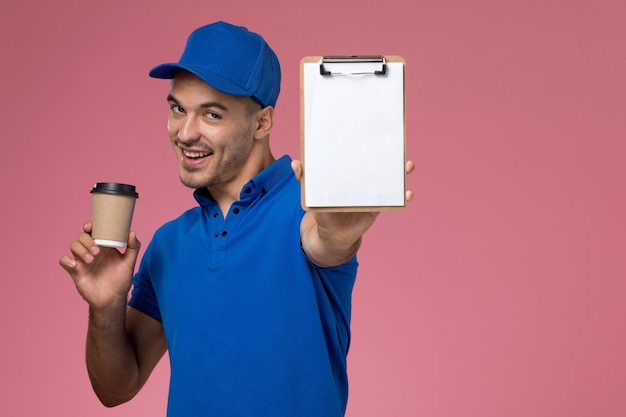 Mensajero masculino en uniforme azul sosteniendo el bloc de notas de la taza de café marrón en rosa, entrega de trabajo de servicio uniforme