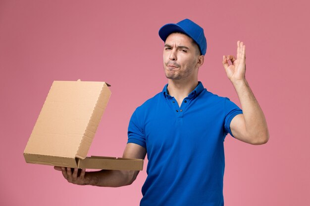 Mensajero masculino en uniforme azul sosteniendo la caja de comida abriéndola en rosa, entrega de trabajo de servicio uniforme