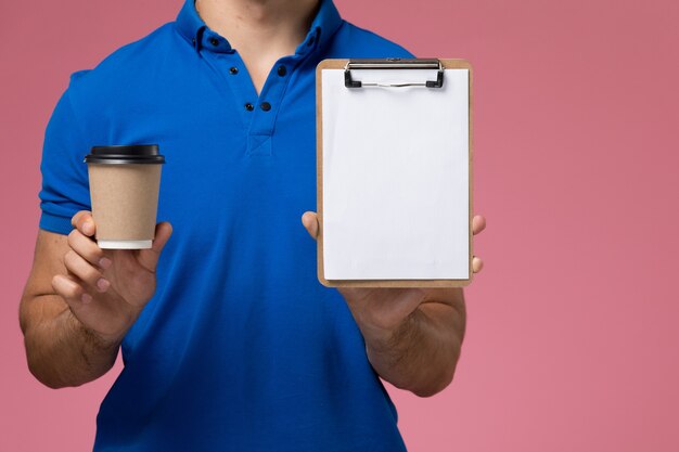 Mensajero masculino en uniforme azul sosteniendo la taza de café y el bloc de notas en rosa, entrega de trabajo de servicio uniforme