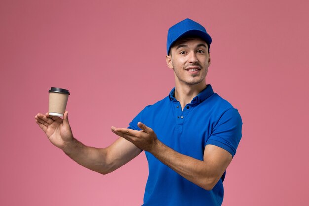 Mensajero masculino en uniforme azul sosteniendo la taza de café posando en rosa, entrega de servicio uniforme de trabajador