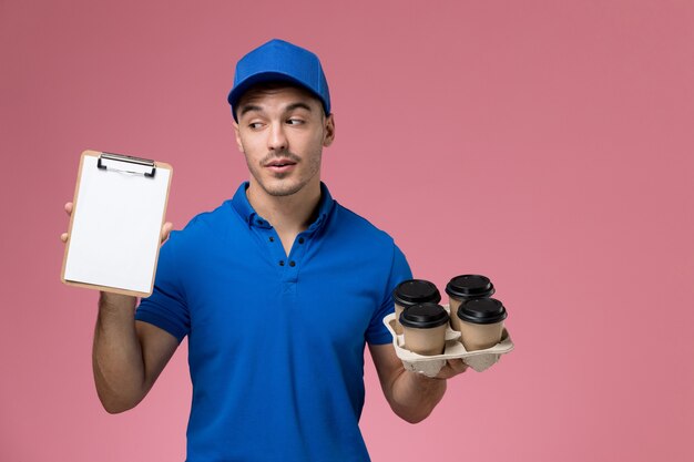 Mensajero masculino en uniforme azul sosteniendo tazas de café de entrega con bloc de notas en rosa, servicio de trabajador uniforme de entrega