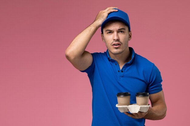 Mensajero masculino en uniforme azul sosteniendo tazas de café de entrega en el piso rosa entrega de trabajo de servicio uniforme