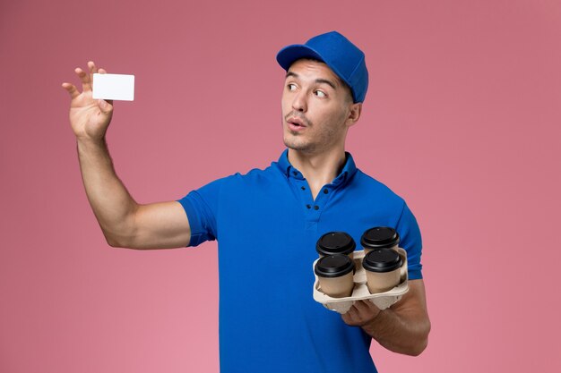 Mensajero masculino en uniforme azul sosteniendo tazas de café de entrega y tarjeta en rosa, servicio de trabajador de trabajo uniforme entrega