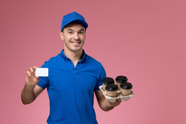 Mensajero masculino en uniforme azul sosteniendo tazas de café de entrega y tarjeta sonriendo en rosa, servicio uniforme entrega de trabajo