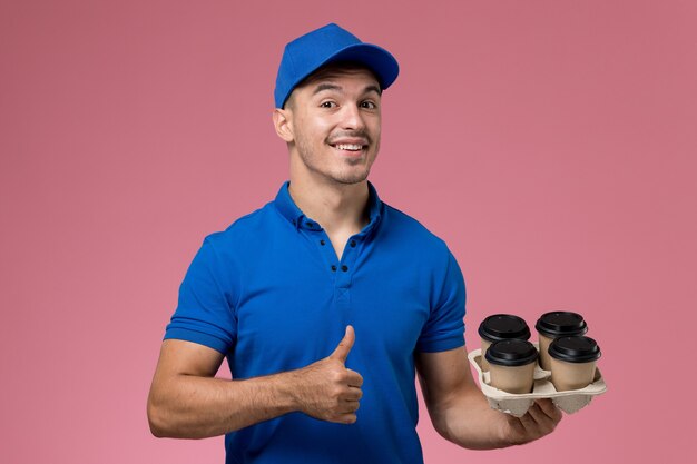 Mensajero masculino en uniforme azul sosteniendo tazas de café con una leve sonrisa en rosa, entrega de servicio uniforme de trabajador