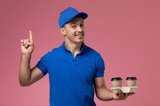 Mensajero masculino en uniforme azul sosteniendo tazas de café marrón en rosa, entrega de trabajo de servicio uniforme