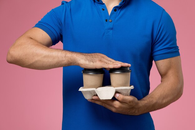 Mensajero masculino en uniforme azul sosteniendo tazas de café marrón sobre rosa, entrega de servicio uniforme de trabajador