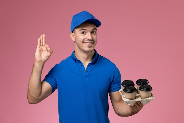 Mensajero masculino en uniforme azul sosteniendo tazas de café y posando en rosa, entrega de servicio uniforme de trabajador