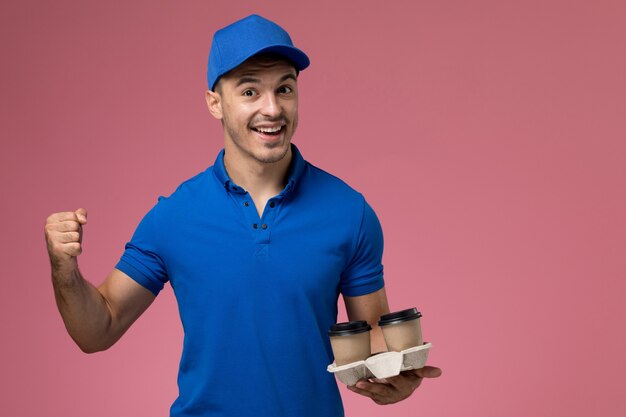 Mensajero masculino en uniforme azul sosteniendo tazas de café regocijándose en rosa, entrega de servicio uniforme de trabajador de trabajo