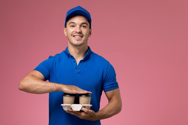 Mensajero masculino en uniforme azul sosteniendo tazas de café y sonriendo en rosa, entrega de trabajo de servicio uniforme