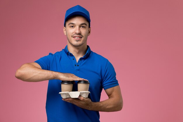 Mensajero masculino en uniforme azul sosteniendo tazas de café y sonriendo en rosa, entrega de trabajo de servicio uniforme