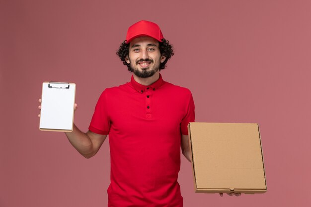 Mensajero masculino de vista frontal en camisa roja y capa con caja de comida de entrega y bloc de notas en la pared rosa