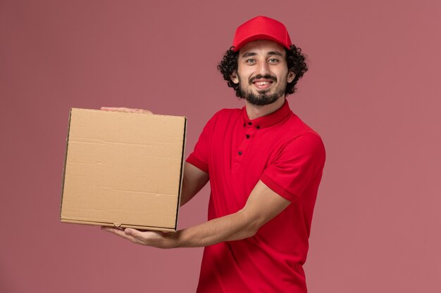 Mensajero masculino de vista frontal en camisa roja y capa con caja de comida de entrega en la pared rosa claro
