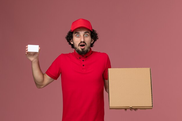 Mensajero masculino de vista frontal en camisa roja y capa con caja de comida de entrega y tarjeta en la pared rosa