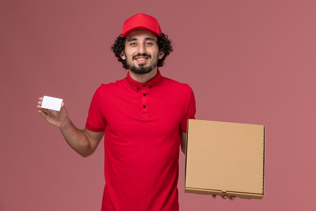Mensajero masculino de vista frontal en camisa roja y capa con caja de comida de entrega y tarjeta en la pared rosa