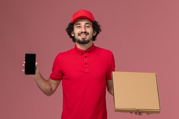 Mensajero masculino de vista frontal en camisa roja y capa con caja de comida de entrega vacía con teléfono en pared rosa