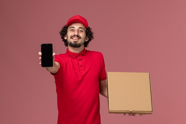 Mensajero masculino de vista frontal en camisa roja y capa con caja de comida de entrega vacía con teléfono en pared rosa