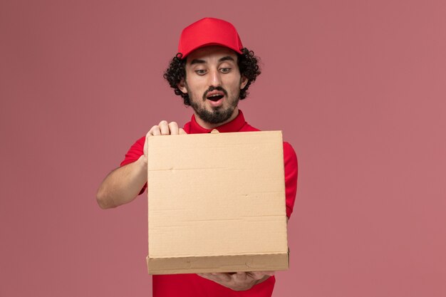 Mensajero masculino de vista frontal con camisa roja y capa sosteniendo la caja de comida de entrega y abriéndola en la pared rosa claro