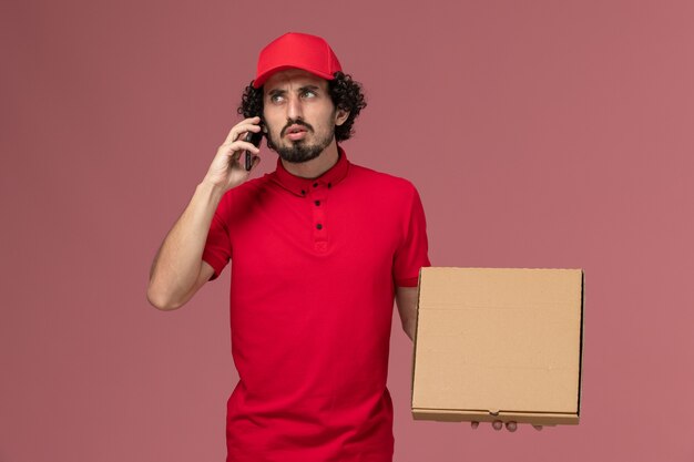 Mensajero masculino de vista frontal con camisa roja y capa sosteniendo una caja de comida de entrega vacía mientras habla por teléfono en la empresa de uniforme de entrega de servicio de escritorio rosa