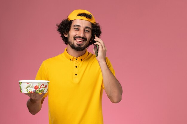 Mensajero masculino de vista frontal en capa uniforme amarilla con cuenco de entrega en sus manos mientras habla por teléfono sobre fondo rosa claro.