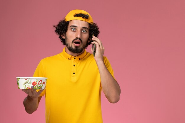Mensajero masculino de vista frontal en capa uniforme amarilla con cuenco de entrega en sus manos mientras habla por teléfono sobre fondo rosa claro.