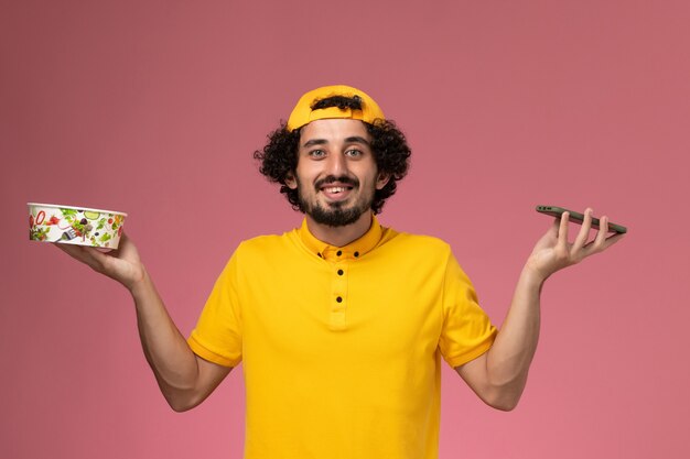 Mensajero masculino de vista frontal en capa uniforme amarilla con tazón de entrega y teléfono inteligente en sus manos sobre fondo rosa claro.