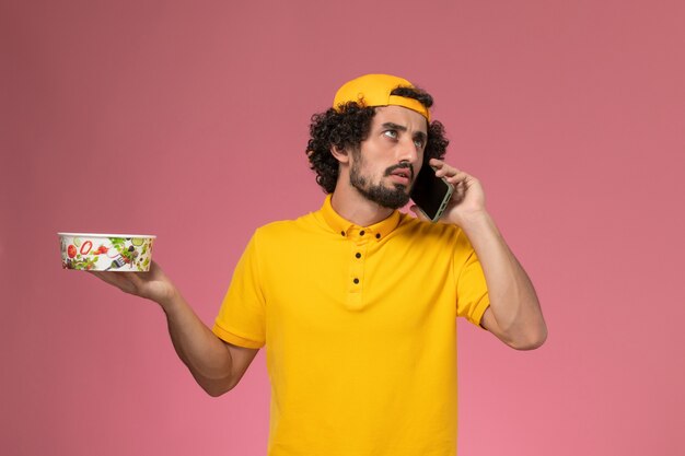 Mensajero masculino de vista frontal en capa uniforme amarilla con tazón de entrega y teléfono inteligente en sus manos sobre fondo rosa claro.