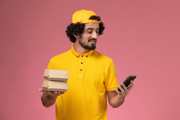 Mensajero masculino de vista frontal en capa uniforme amarilla con teléfono y paquetes de alimentos en sus manos sobre fondo rosa claro.