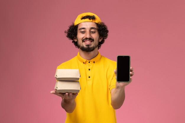 Mensajero masculino de vista frontal en capa uniforme amarilla con teléfono y paquetes de alimentos en sus manos sobre fondo rosa claro.