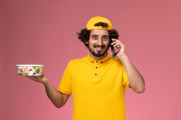 Mensajero masculino de vista frontal en capa uniforme amarillo con cuenco de entrega en sus manos hablando por teléfono sobre el fondo rosa claro.
