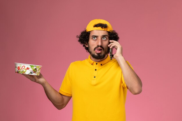 Mensajero masculino de vista frontal en capa uniforme amarillo con cuenco de entrega en sus manos hablando por teléfono sobre el fondo rosa claro.