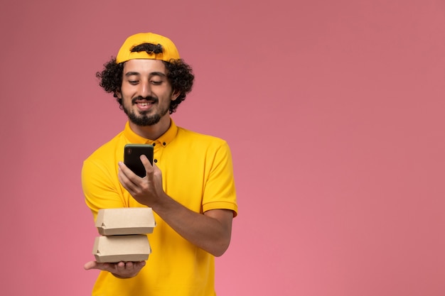 Mensajero masculino de vista frontal en capa uniforme amarillo con paquetes de alimentos en sus manos hablando por teléfono sobre fondo rosa claro.