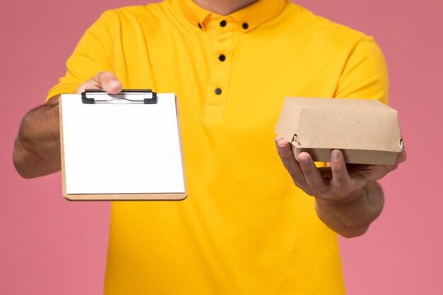 Mensajero masculino de vista frontal cercana en uniforme amarillo y gorra con bloc de notas y pequeño paquete de comida de entrega en sus manos en la pared rosa claro