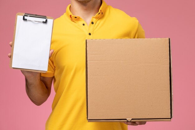 Mensajero masculino de la vista frontal cercana en uniforme amarillo sosteniendo el bloc de notas y la caja de entrega de alimentos sobre fondo rosa