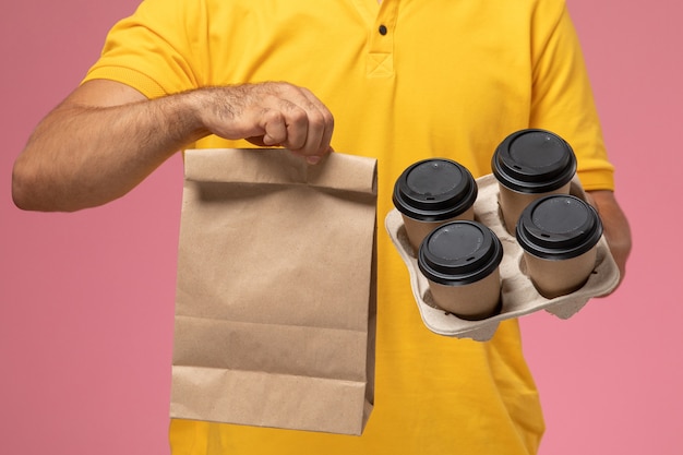 Mensajero masculino de vista frontal cercana en uniforme amarillo sosteniendo el paquete de alimentos y tazas de café de entrega sobre fondo rosa