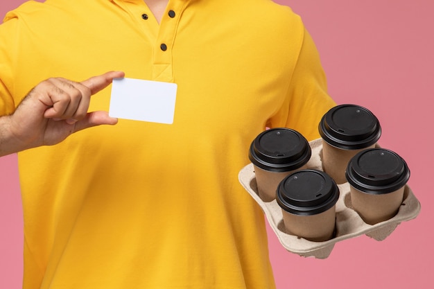 Mensajero masculino de vista frontal cercana en uniforme amarillo con tarjeta de plástico y tazas de café de entrega sobre fondo rosa