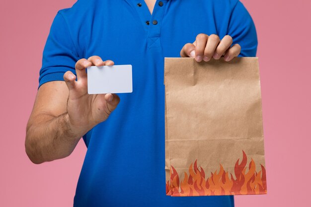 Mensajero masculino de vista frontal cercana en uniforme azul con paquete de alimentos y tarjeta en la entrega de trabajo de servicio uniforme rosa