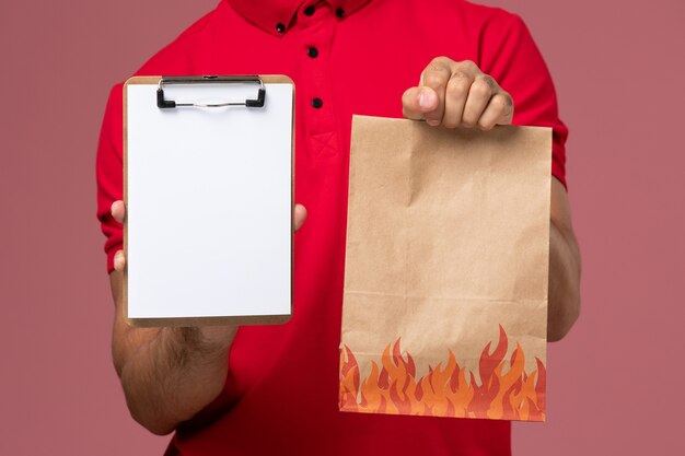 Mensajero masculino de vista frontal cercana en uniforme rojo y capa con paquete de alimentos y bloc de notas en uniforme de trabajo de trabajador de entrega de servicio de pared rosa