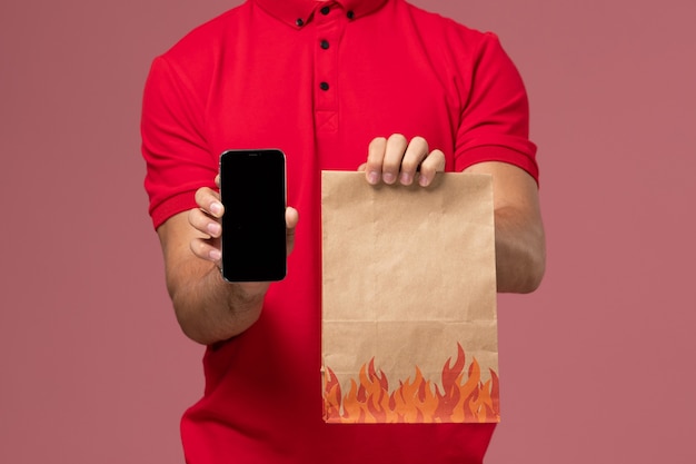 Mensajero masculino de vista frontal cercana en uniforme rojo y capa sosteniendo el paquete de alimentos y el teléfono en la pared rosa