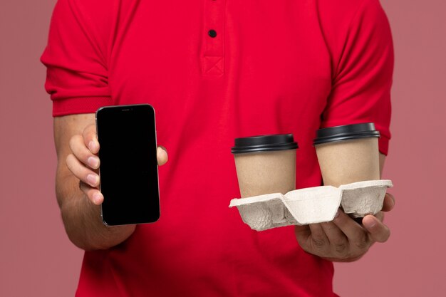 Mensajero masculino de vista frontal cercana en uniforme rojo sosteniendo tazas de café de entrega marrón y teléfono en la pared rosa claro