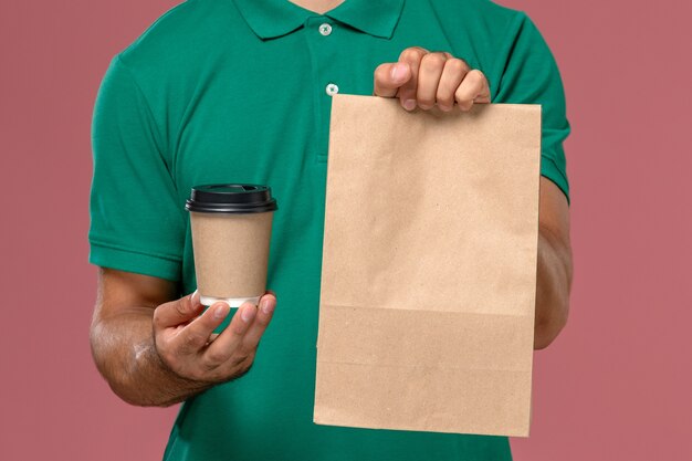 Mensajero masculino de la vista frontal cercana en uniforme verde sosteniendo la taza de café de entrega y el paquete de alimentos sobre fondo rosa claro