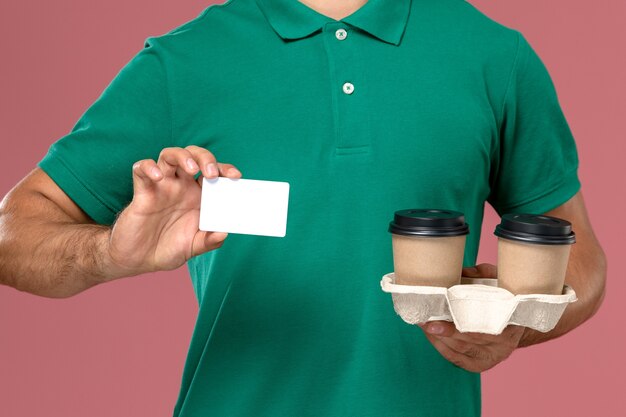 Mensajero masculino de vista frontal cercana en uniforme verde sosteniendo tazas de café de entrega marrón y tarjeta de plástico blanco sobre fondo de escritorio rosa