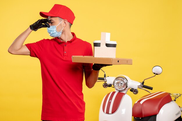 Mensajero masculino de vista frontal en máscara con entrega de café y caja en trabajo amarillo servicio de color covid- virus uniforme trabajo pandémico