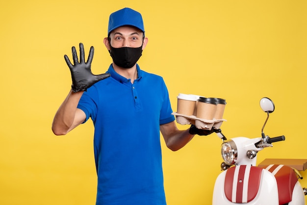 Mensajero masculino de vista frontal en máscara negra sosteniendo café en trabajo amarillo covid- trabajo uniforme de entrega pandémica