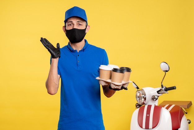 Foto gratuita mensajero masculino de vista frontal en máscara negra sosteniendo café en trabajo amarillo covid- trabajo uniforme de servicio pandémico