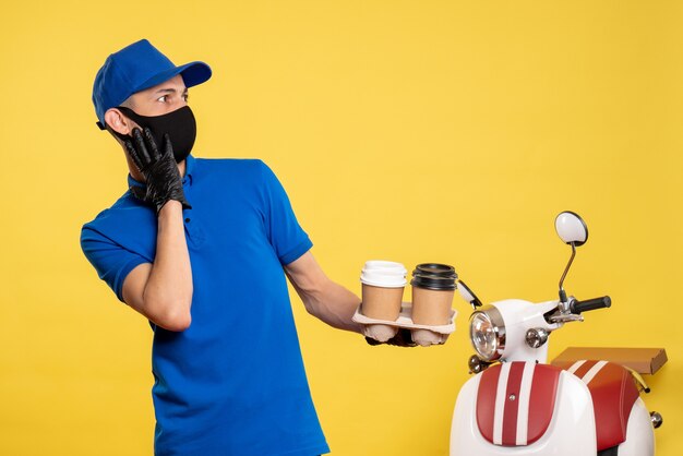 Mensajero masculino de vista frontal con máscara negra sosteniendo café en el trabajo amarillo covid- uniforme de servicio de trabajo pandémico