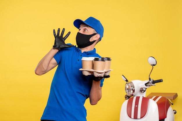 Mensajero masculino de vista frontal en máscara negra sosteniendo café en un trabajo de entrega de trabajo amarillo uniforme de servicio de pandemia covid