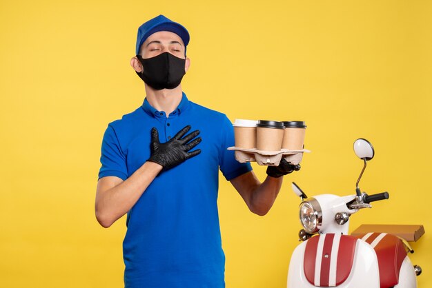 Mensajero masculino de vista frontal en máscara sosteniendo café en trabajo amarillo covid- trabajo de entrega de servicios pandémicos