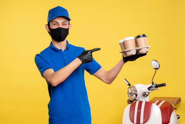 Mensajero masculino de vista frontal en máscara sosteniendo café en uniforme de trabajo amarillo covid- servicio de pandemia de entrega