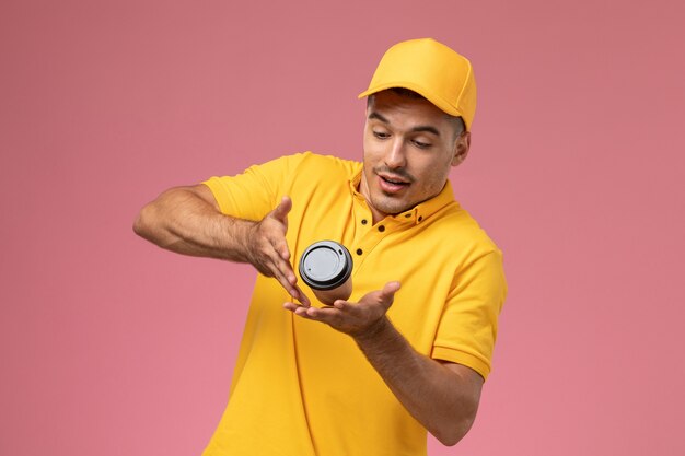 Mensajero masculino de vista frontal en uniforme amarillo apenas sosteniendo la taza de entrega de café marrón sobre el fondo rosa